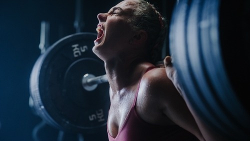 Athletic woman shouting with effort while performing a heavy barbell back squat in a gym.