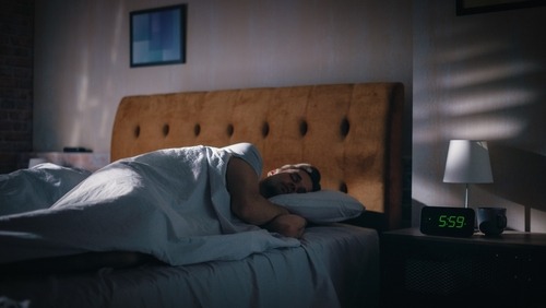 A man sleeping peacefully in a dimly lit bedroom with a digital alarm clock showing 5:59 AM.