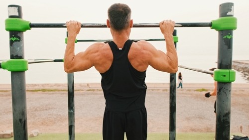 Muscular man performing pull-ups at an outdoor calisthenics gym on the beach.