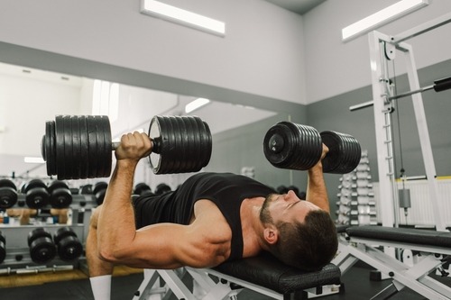 A muscular man performing a chest press with heavy dumbbells on a flat bench in a modern gym.