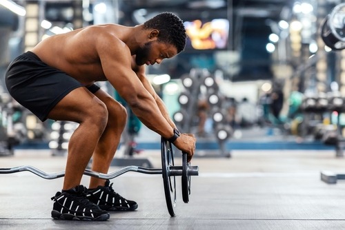 Shirtless athletic man preparing to lift an EZ curl barbell during a strength training workout in a gym.