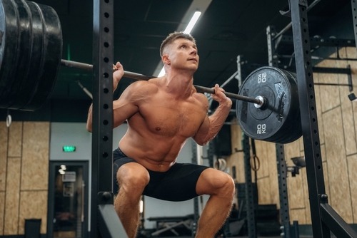 Muscular shirtless man performing a heavy barbell back squat in a gym power rack.