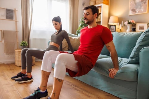 A man and woman performing tricep dips using a sofa during a home workout session.