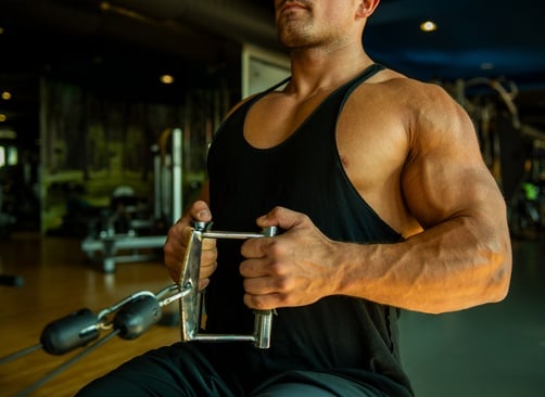 Muscular man in a black tank top performing seated cable rows for back strength training at a gym.