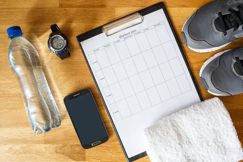 Fitness workout plan on a clipboard with running shoes, water bottle, and gym accessories on a wooden floor.