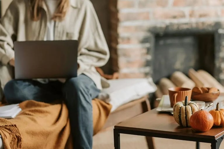 woman on laptop in her cozy fall nook with coffee, pumpkins and golden throw with fireplace
