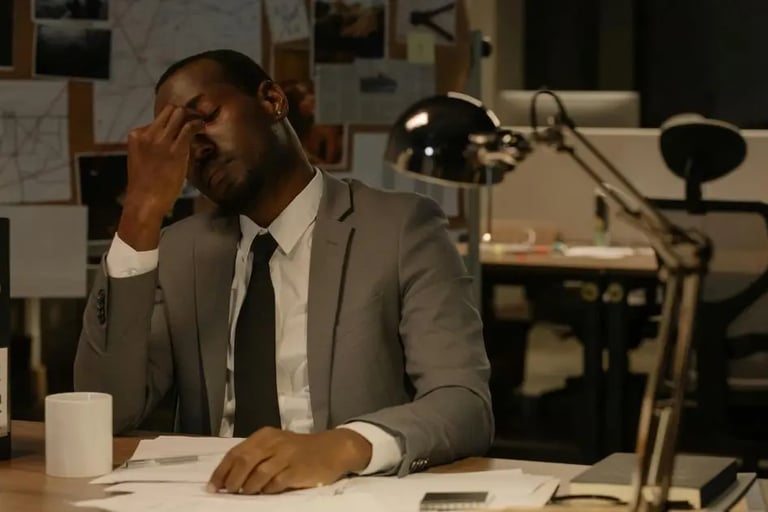A tired man sitting behind a desk piled with paperwork, looking overwhelmed and exhausted, symbolizing the challenges of work