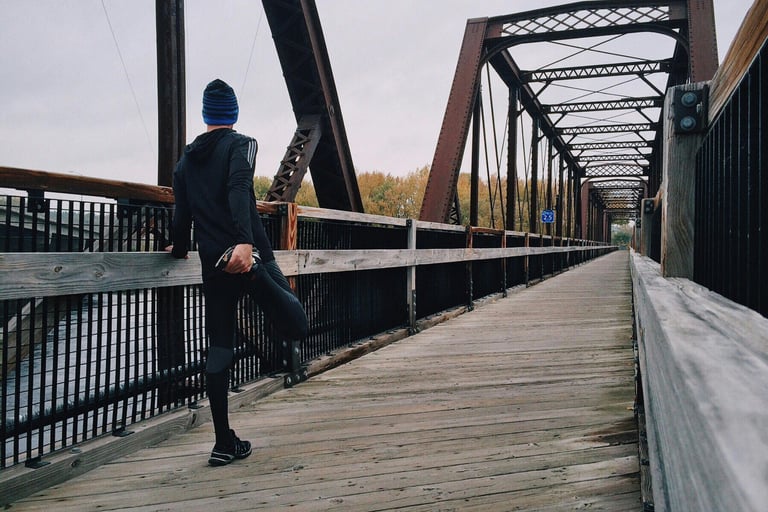 a man in a blue hat and a blue jacket is standing on a bridge. he is warming up for a run to maintai