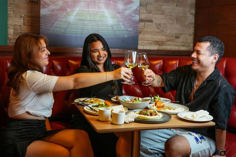Three friends are sitting at a table in a restaurant, enjoying a meal and drinks. They are smiling.