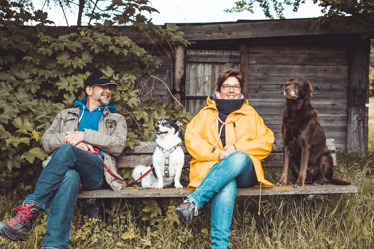 Marion und Martin Lindhof auf einer Bank im Wald, vor einer Holzhütte mit brauner Labradorhündin Amy