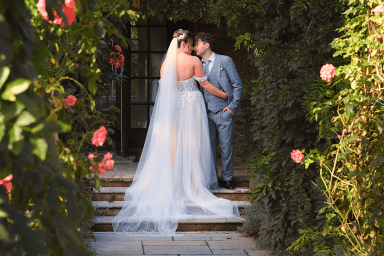 A bride and groom embracing at the end of a path, surrounded by flowers and greenery.