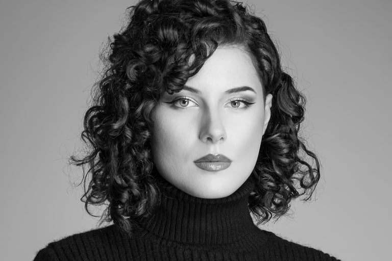  Ein Schwarz-Weiß-Porträt einer Frau mit kurzen, lockigen Haaren. Sie trägt einen dunklen Rollkragenpullover und hat einen au