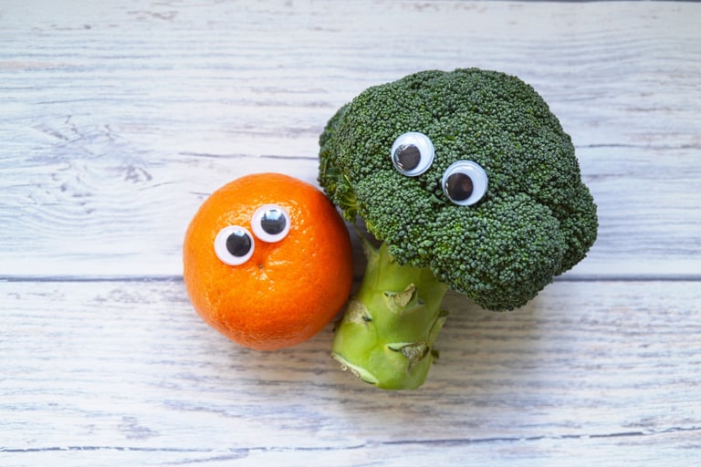 a broccoli and an orange with plastic eyes are cuddled up together on a table