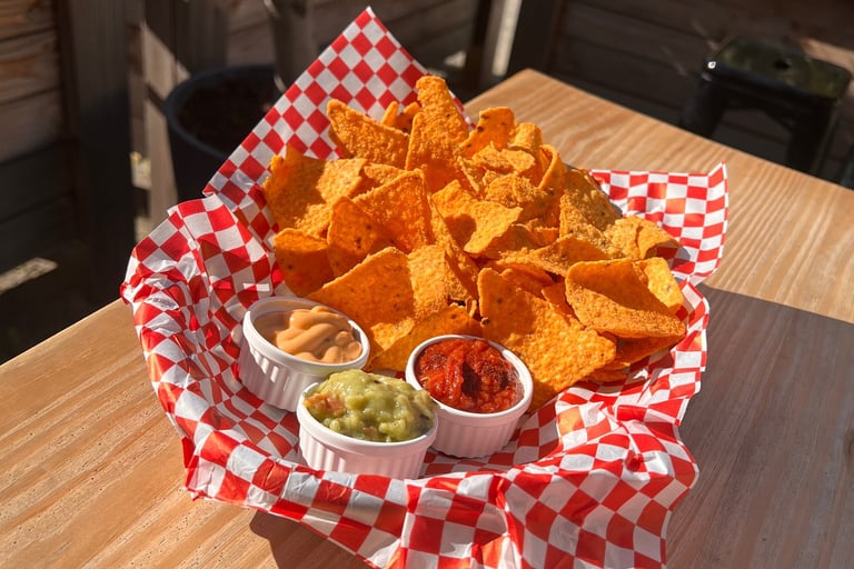 A basket of crunchy tortilla chips served with salsa, guacamole, and cheese dip on an outdoor patio table.
