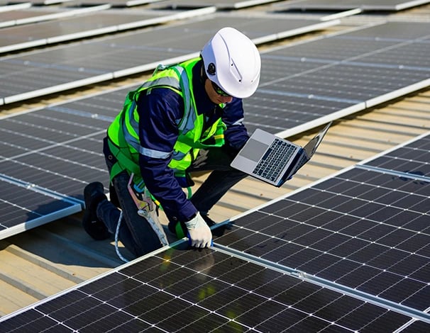 Engineer in safety gear using a laptop to inspect solar panel installation on a rooftop.