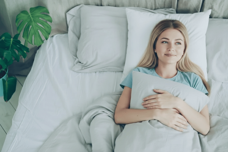 A smiling young woman lying in a comfortable bed with white pillows and a monstera plant.