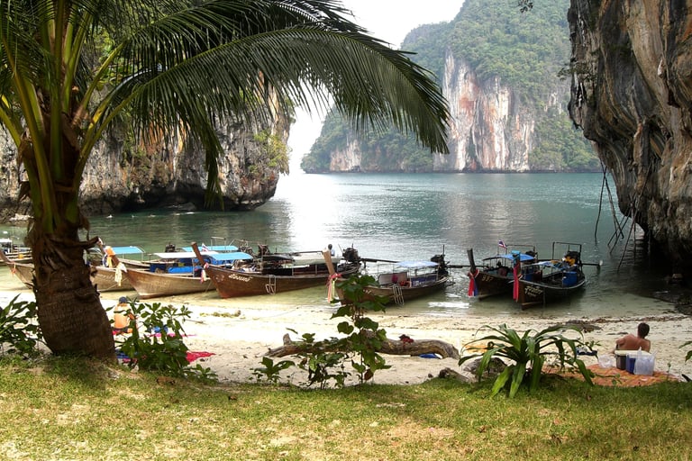 Traditional Thai longtail boats anchored at a tropical beach with limestone cliffs and palm trees