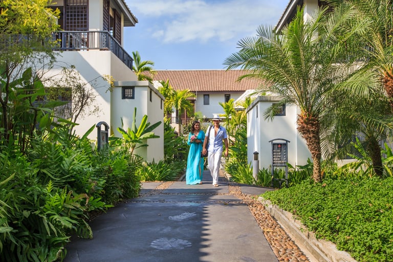 A couple walking down a tropical resort pathway lined with lush greenery, ferns, and palm trees