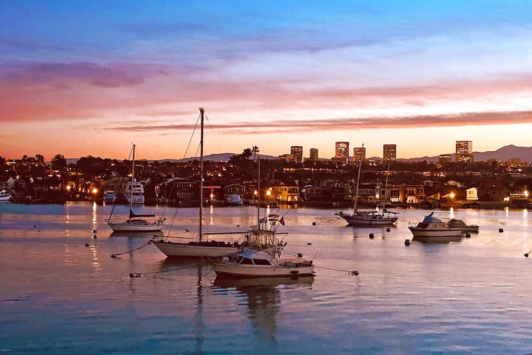 Newport Harbor before dawn, distant city lights glowing across still water.
