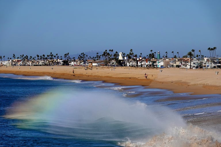 A breaking wave at Balboa Pier creating a fleeting rainbow in the spray, palm trees in the background.