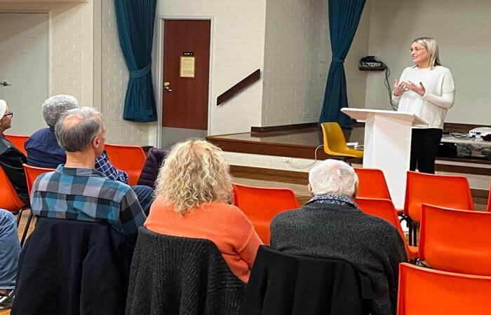 Candice speaks at a podium to a small seated audience in a community hall during a church gathering.