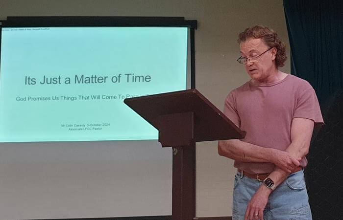 Collin stands at a lectern reading from notes while a presentation titled “It’s Just a Matter of Tim
