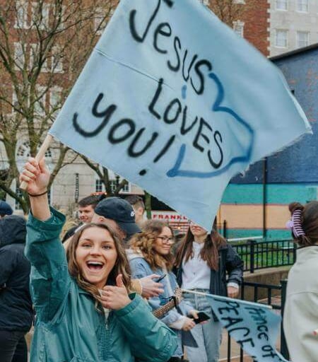 Smiling person holding a light blue flag that reads “Jesus LOVES you!!” during a public gathering.