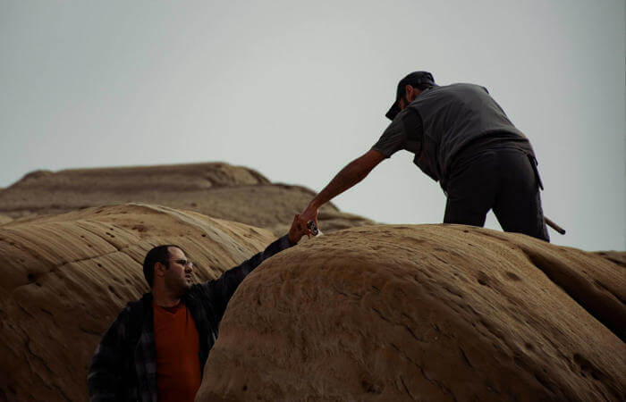 One person reaching down to help another climb a rocky slope under a cloudy sky