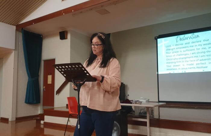 Hazel reads from a stand during a church meeting, with a projected prayer or declaration visible in 