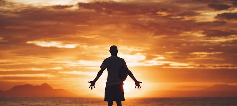 A person stands with open arms at sunset, expressing wonder, surrender, and spiritual searching.