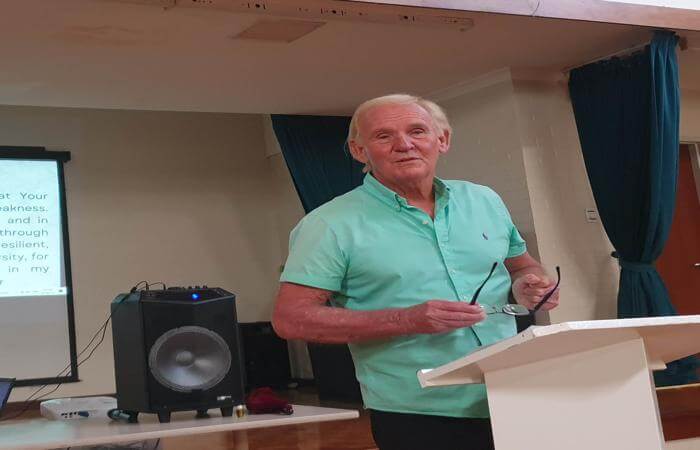 Len stands at a podium holding glasses, smiling as he addresses a church group in a relaxed setting.