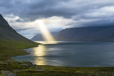 Sunlight breaks through clouds over a peaceful valley and water, showing divine presence in nature.