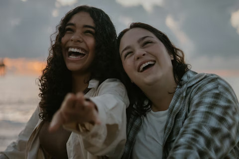 Two friends laugh together by the water, reflecting joy, connection, and transformed lives.