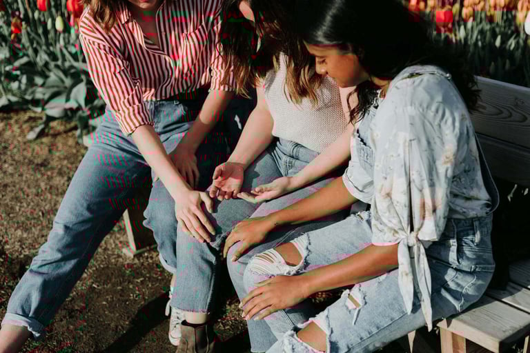 two young women sitting on a bench in a park praying for someone