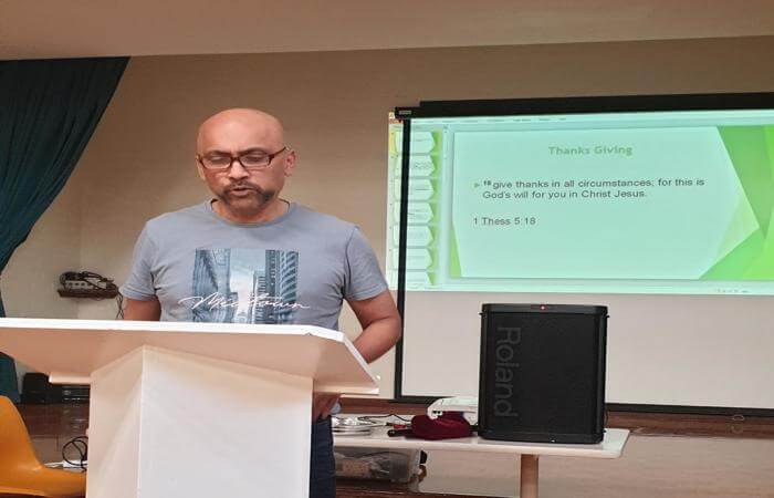 Raju reads from a lectern during a church gathering, with a slide behind him displaying a Bible vers