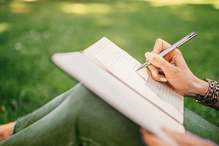 a woman person holding a pen and writing on a notebook outside