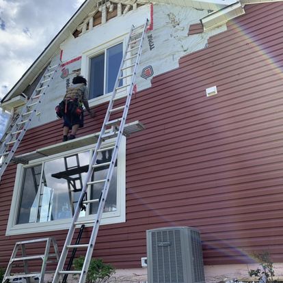 A contractor on a ladder installing red vinyl siding on the exterior of a residential home.