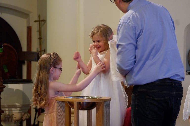 2 young children looking happy at a church font as a Vicar baptises them. 