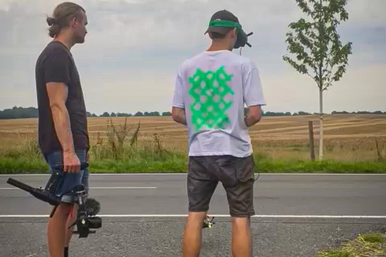 two men standing on a road with a camera and a flying fpv drone following a bmw car