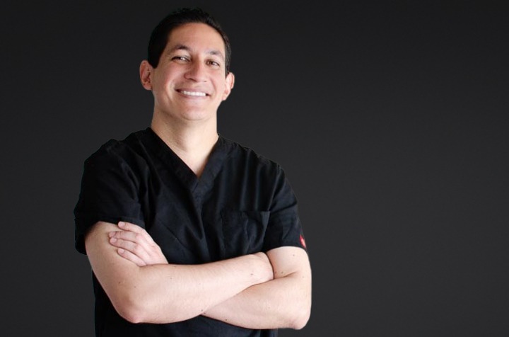 Smiling male healthcare professional in black scrubs with arms crossed against a dark background.