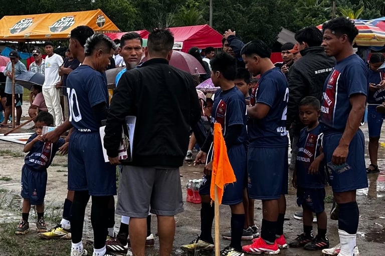 A soccer team in blue uniforms huddles with their coach on a muddy outdoor field during a tournament.