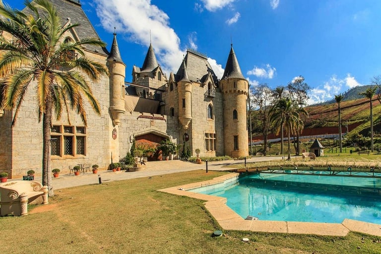 Fachada de pedra do Castelo de Itaipava em Petrópolis com torres e piscina ao sol.