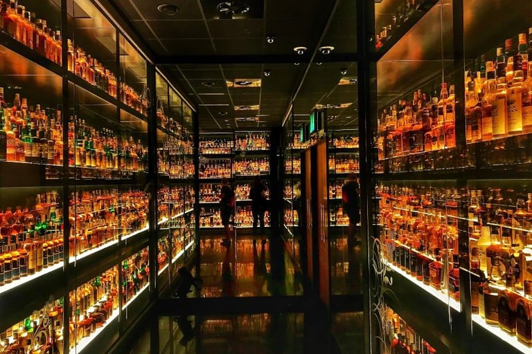 A vast collection of whiskey bottles displayed on illuminated glass shelves in a dark spirits cellar.