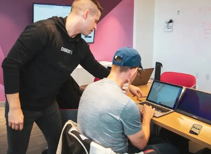Two individuals collaborating in a modern tech workspace with laptops, screen, and whiteboard for UI planning.