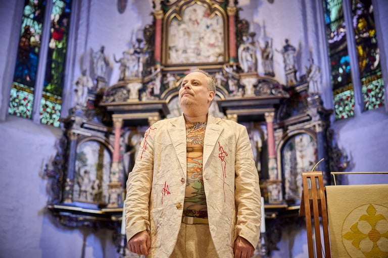 Ein Schauspieler vor einem Kirchenaltar. Er schaut in die Ferne.