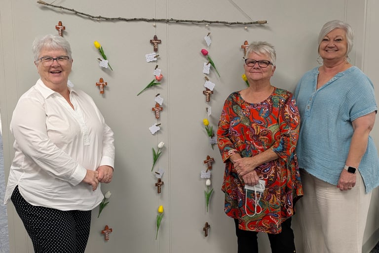 Three smiling women stand beside a hanging prayer wall display with wooden crosses and tulips.