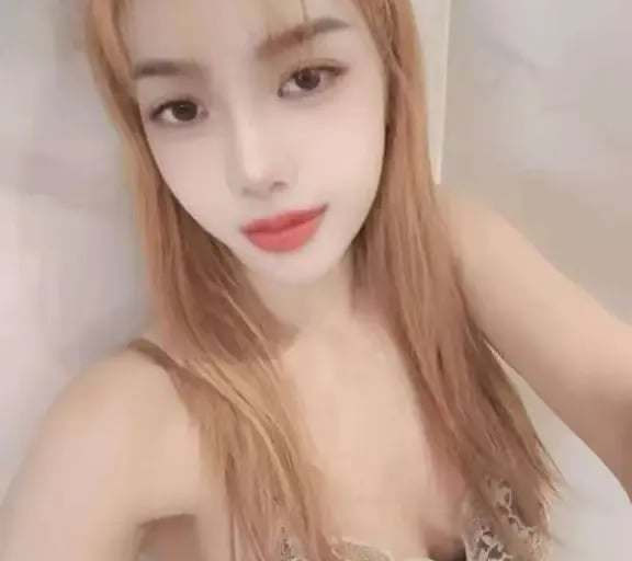 A young woman with long blonde hair wearing a lace-trim black camisole poses for a selfie.