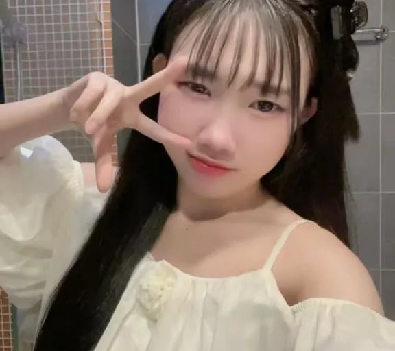 Young woman with long dark hair and bangs wearing a white off-the-shoulder top posing with a peace sign.