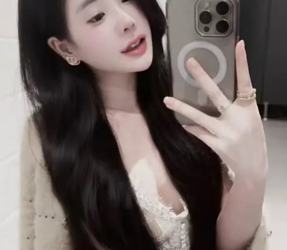 Young woman with long dark hair taking a mirror selfie wearing a lace top and fuzzy cardigan.