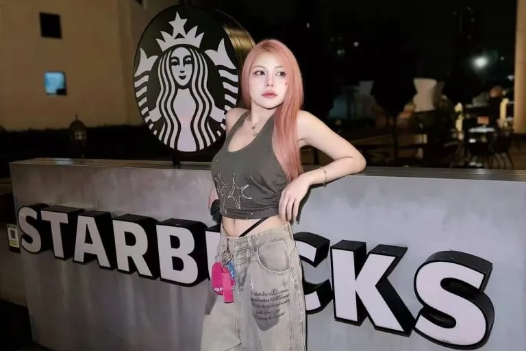 A stylish woman with pink hair posing outside a Starbucks cafe at night in trendy streetwear.